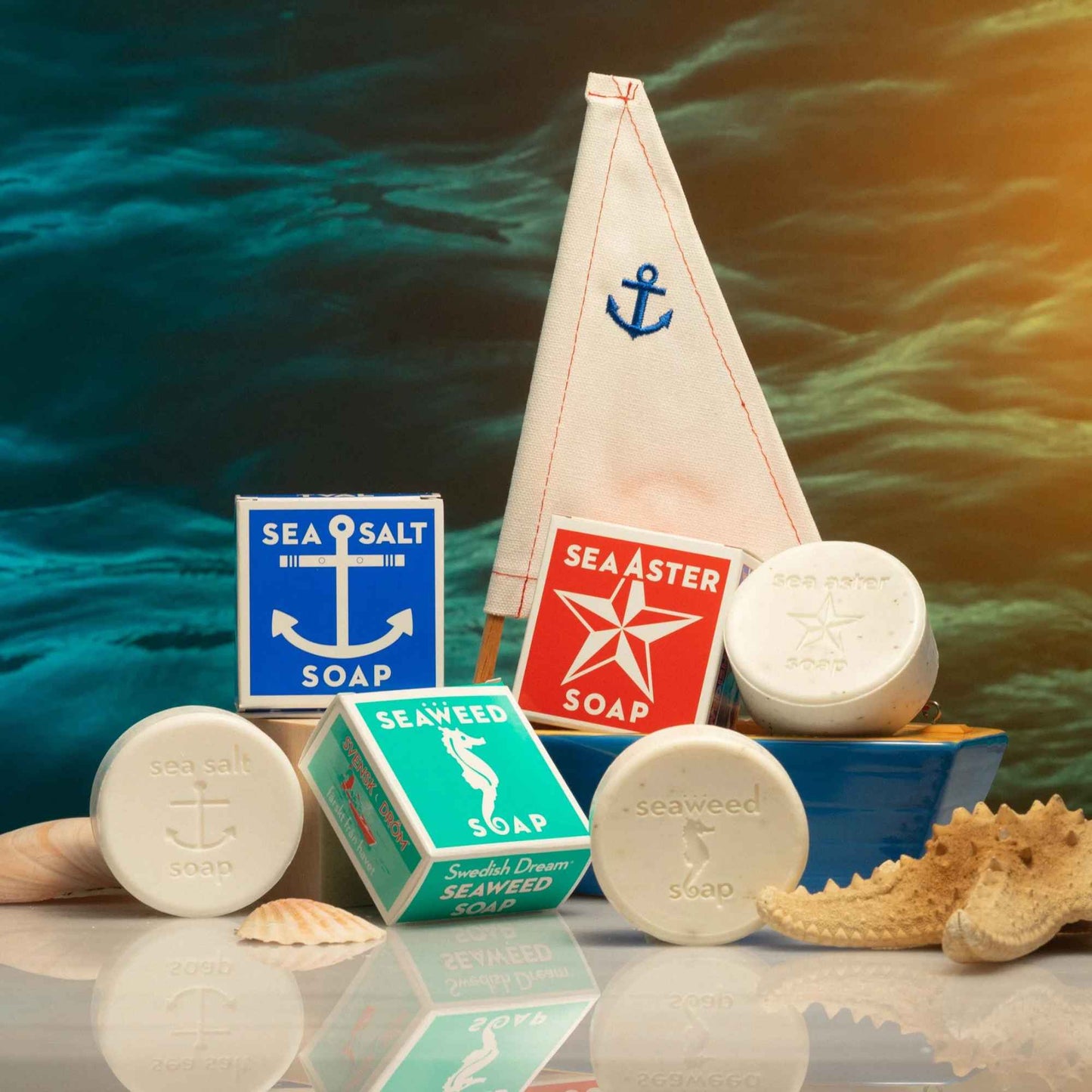 Front view of Swedish Dream Sea Salt Soap with sailboat packaging, soap bars, and seashells against a watery background