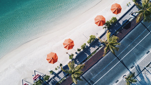 Aerial view of a palm-lined beach road with orange umbrellas beside turquoise water