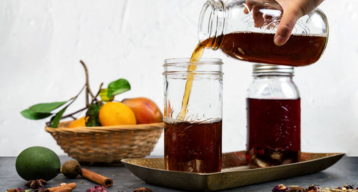 A hand pours herbal cold brew tea into a glass jar beside citrus, spices, and another jar on a tray