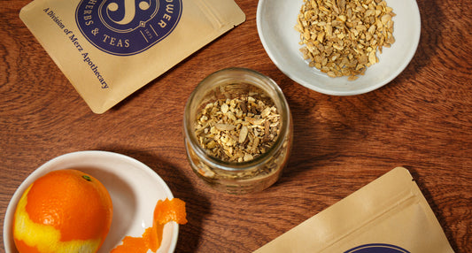 Ingredients for maple orange bitters arranged with orange peel, dried botanicals, and a jar on a wooden table