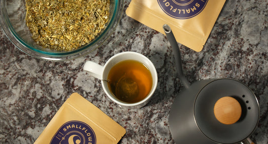 Cup of herbal tea with loose herbs, tea packages, and a teapot on a stone countertop