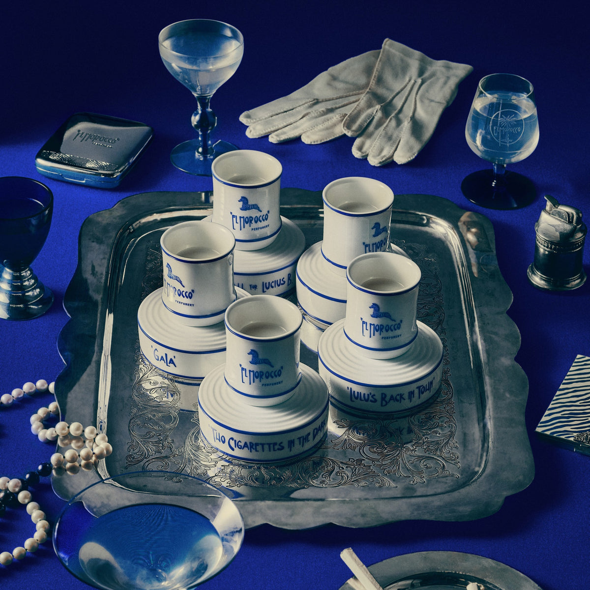A styled flat-lay of a vintage collection featuring a silver tray holding stacked white and blue 'J. Mitchell' cigar boxes, white gloves, blue glass goblets, a pearl necklace, and a small silver pocket watch on a deep blue background.