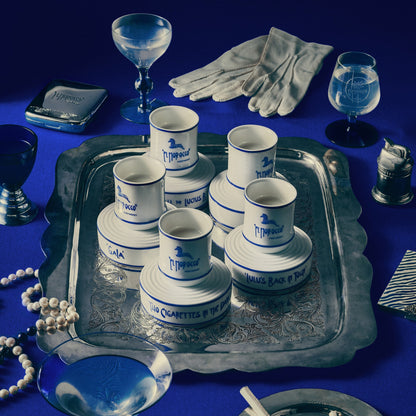 A styled flat-lay of a vintage collection featuring a silver tray holding stacked white and blue 'J. Mitchell' cigar boxes, white gloves, blue glass goblets, a pearl necklace, and a small silver pocket watch on a deep blue background.