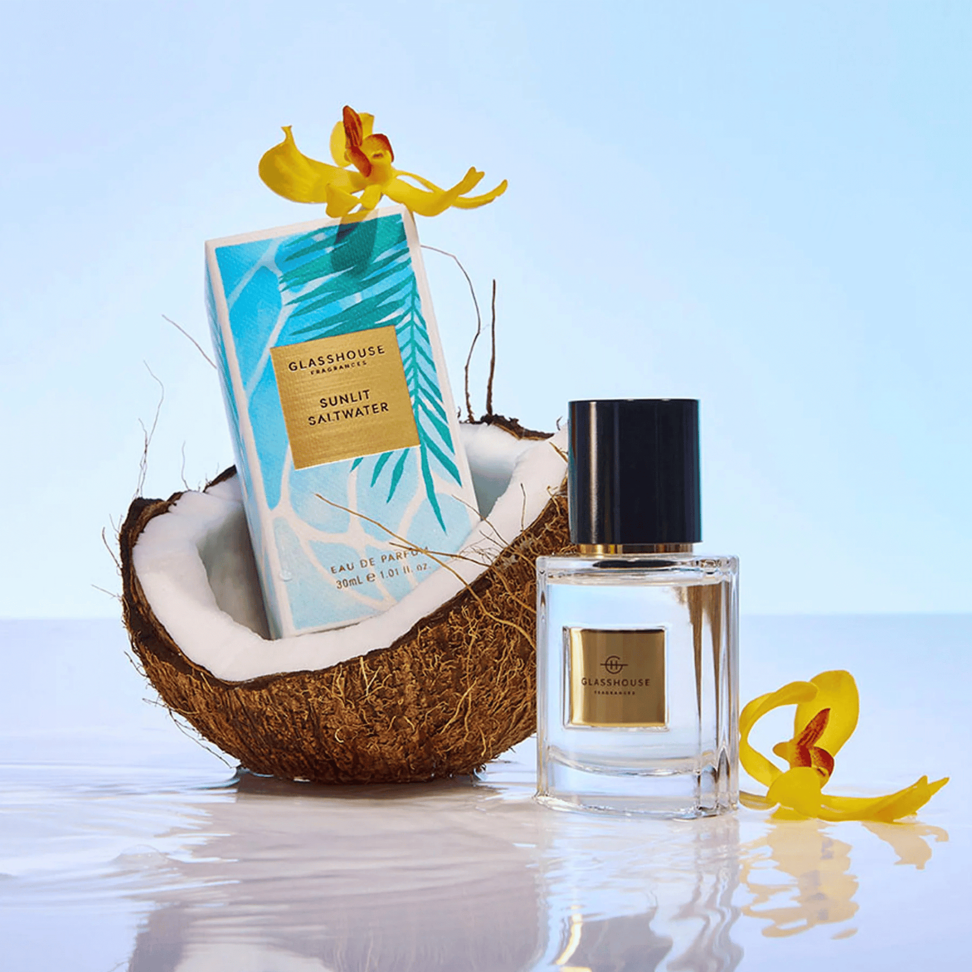 Alternate image of GlassHouse Sunlit Saltwater EDP bottle and packaging in a coconut shell along with tropical flowers 