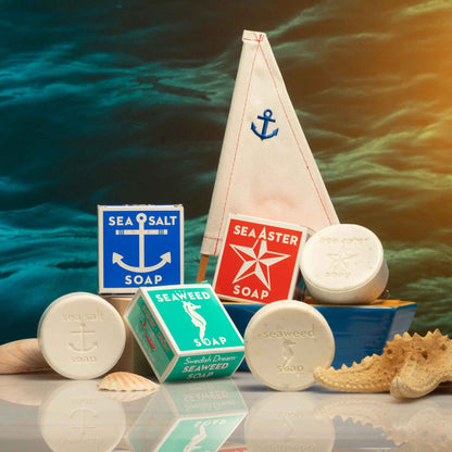 Front view of Swedish Dream Sea Salt Soap with sailboat packaging, soap bars, and seashells against a watery background